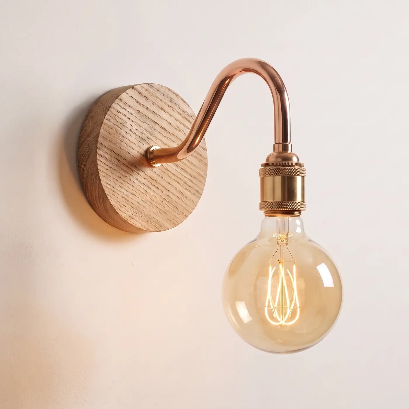 Swan neck wall light with a natural oak back plate, copper swan neck arm and brass bulb holder, lit with a globe filament bulb